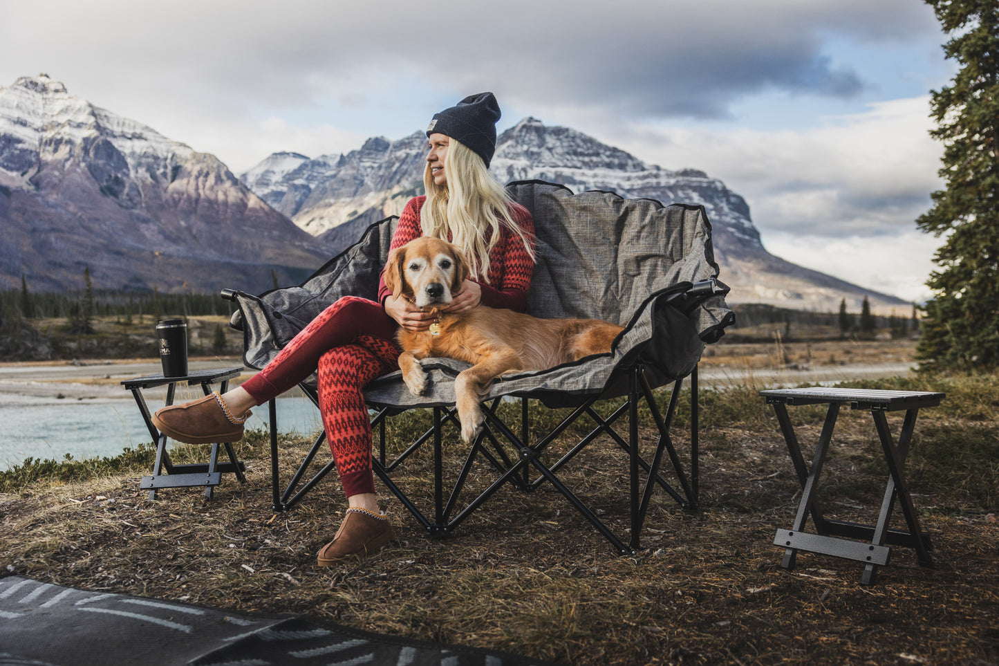 Person sitting on a camping chair with a dog, surrounded by mountains and nature.