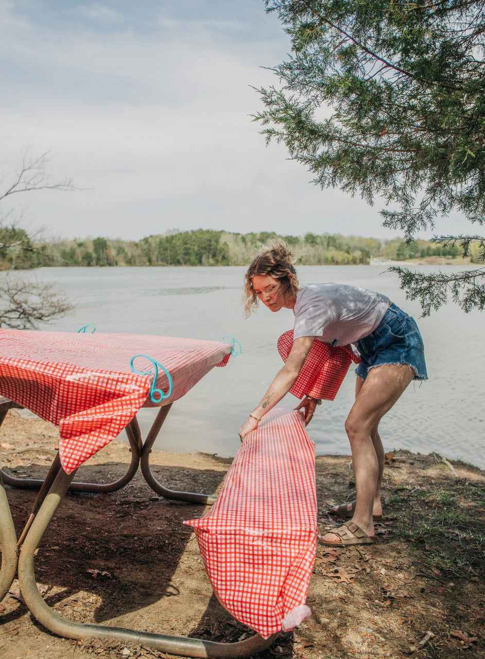 A woman outdoors by a lake sets up a red and white gingham picnic tablecloth on a metal picnic table, with trees and water in the background.