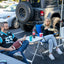 Two people sitting in folding chairs at a tailgate or outdoor gathering, with a black off-road Jeep Wrangler parked behind them. One person holds a football, and the other holds a plate.