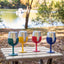 Four insulated wine glasses with colorful silicone sleeves in green, yellow, red, and blue, lined up on a picnic table outdoors with a lake and trees in the background.