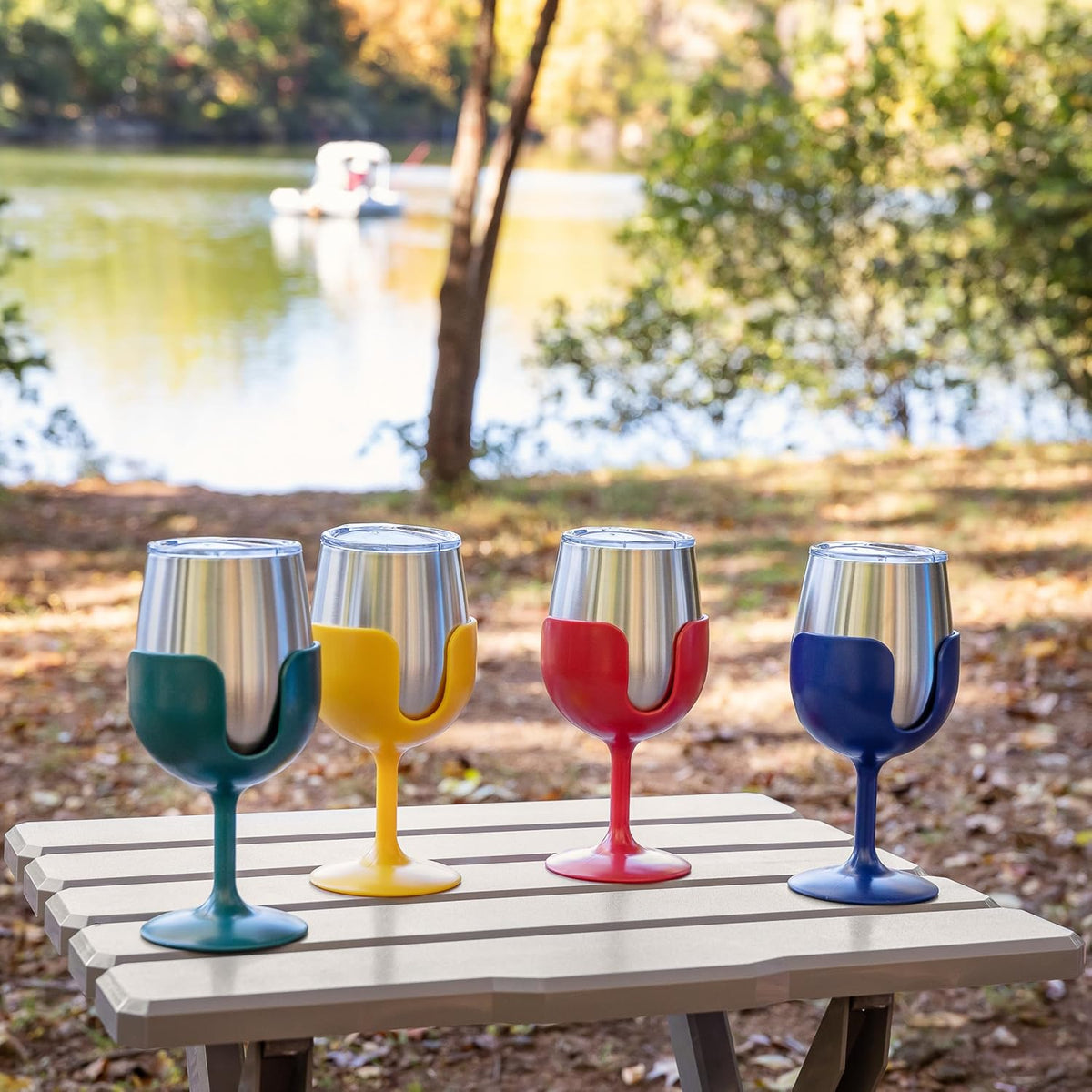 Four insulated wine glasses with colorful silicone sleeves in green, yellow, red, and blue, lined up on a picnic table outdoors with a lake and trees in the background.