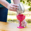 Person holding a silver cup with a pink silicone cup holder on a wooden surface.