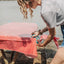 Person arranging a red and white checkered tablecloth on a wooden table outdoors.
