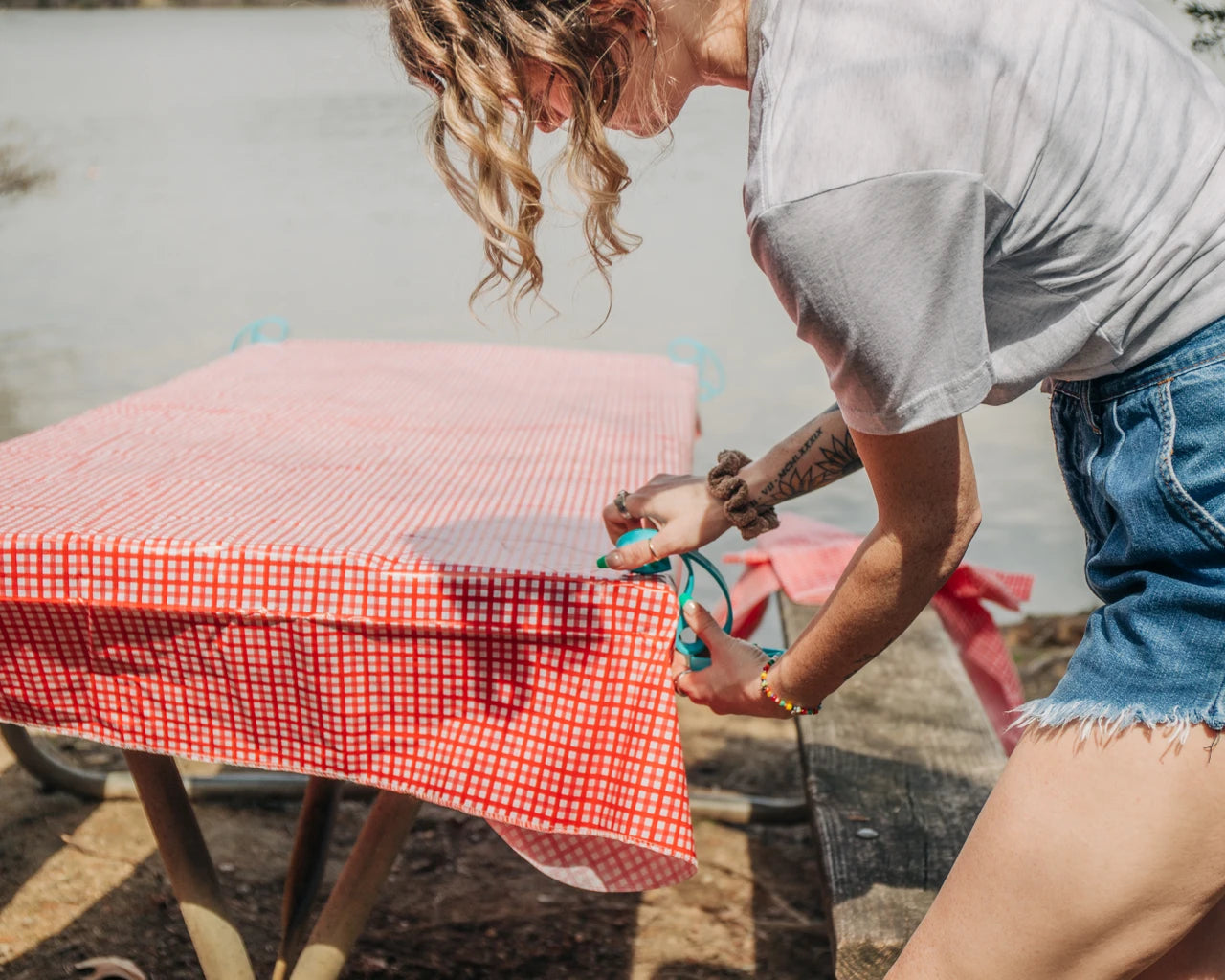 Person arranging a red and white checkered tablecloth on a wooden table outdoors.