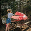 A woman stands at a picnic table in a wooded outdoor setting, holding up a large pink mesh or fabric sheet. Trees, dirt ground, and another picnic table are visible in the background.