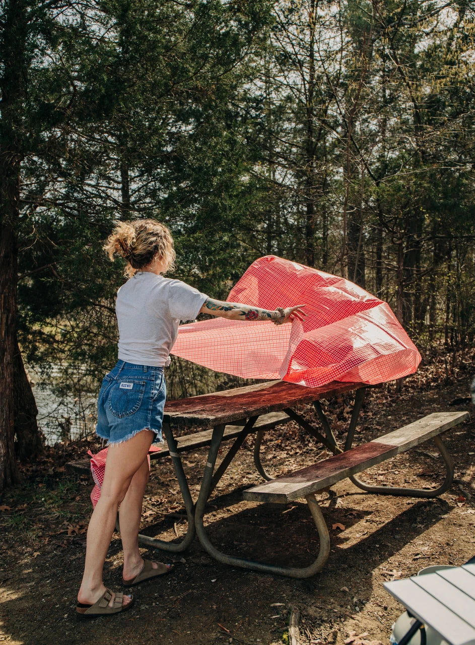 A woman stands at a picnic table in a wooded outdoor setting, holding up a large pink mesh or fabric sheet. Trees, dirt ground, and another picnic table are visible in the background.