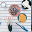 Outdoor picnic-style flat lay on a white slatted table with s'mores ingredients: a plate of Hershey's chocolate bars, marshmallows in a bowl, graham crackers, three metal s'mores grills, a lighter, and a candle.