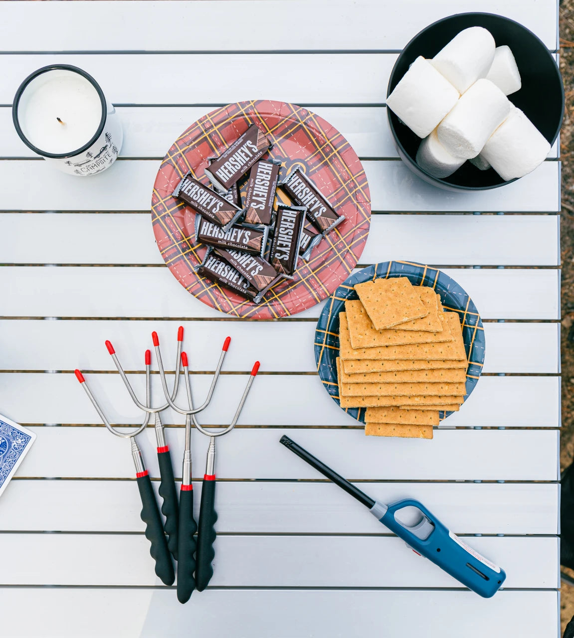 Outdoor picnic-style flat lay on a white slatted table with s'mores ingredients: a plate of Hershey's chocolate bars, marshmallows in a bowl, graham crackers, three metal s'mores grills, a lighter, and a candle.