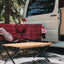 Outdoor camping setup with a red plaid blanket, wooden table, and white van in a natural setting.