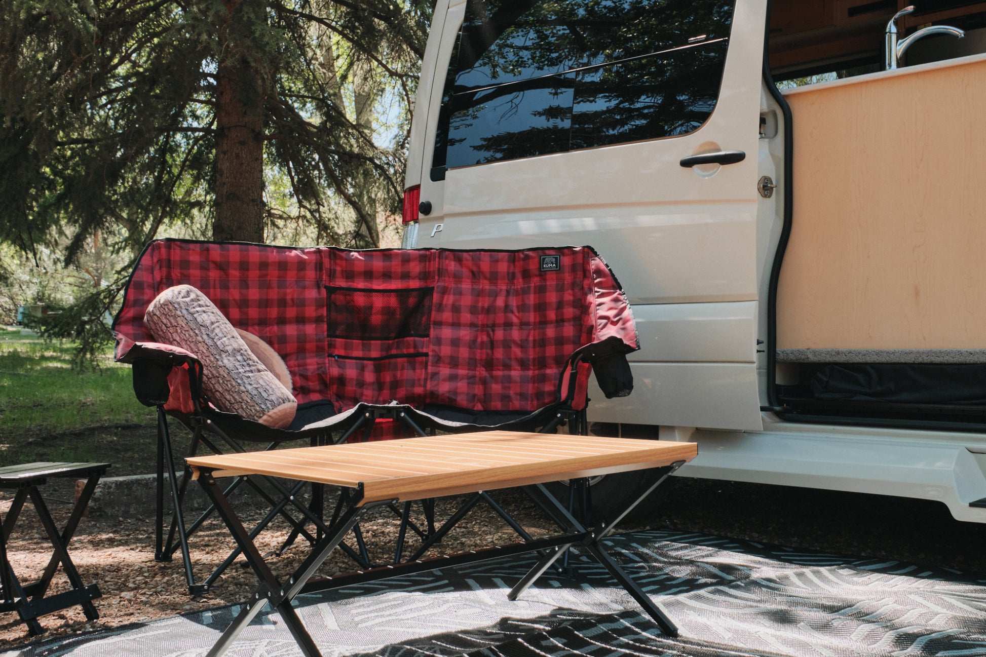 Outdoor camping setup with a red plaid blanket, wooden table, and white van in a natural setting.