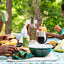 Outdoor picnic scene with people enjoying food and drinks on a table.