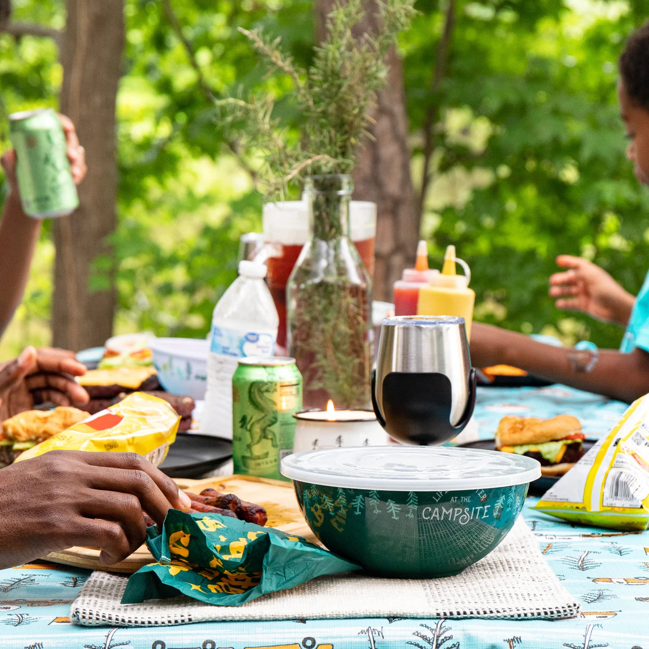 Outdoor picnic scene with people enjoying food and drinks on a table.