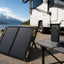 A foldable black solar panel standing on a gravel campsite beside an RV, with a portable power station and smartphone on a table in front of a lake and mountains.