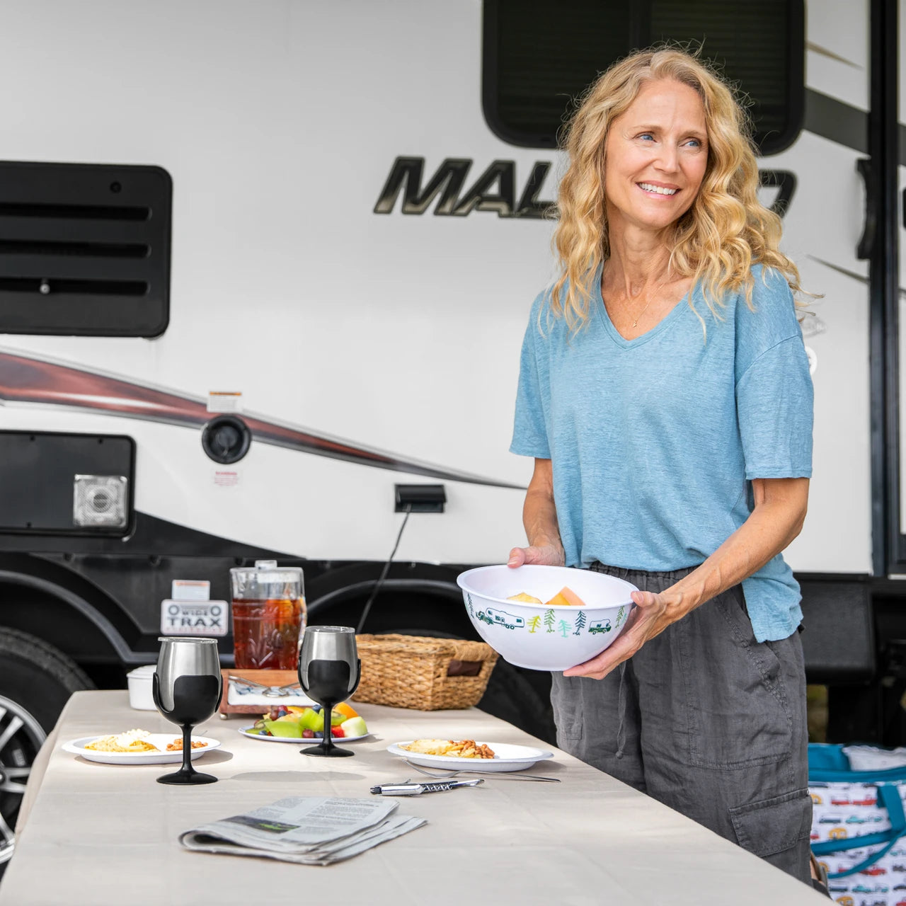 Woman standing by a table with food and drinks in front of an RV, holding a bowl.