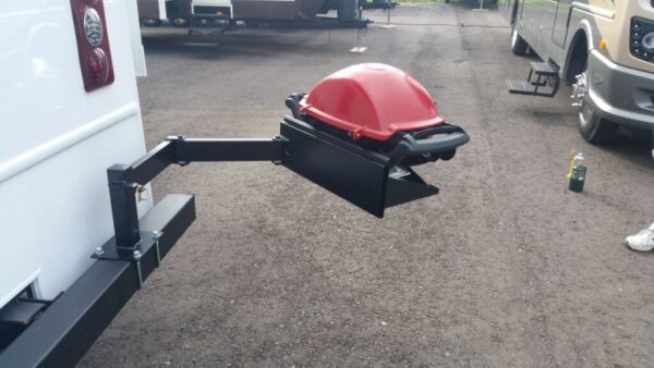 Red grill attached to a vehicle on a gravel surface with other vehicles in the background.