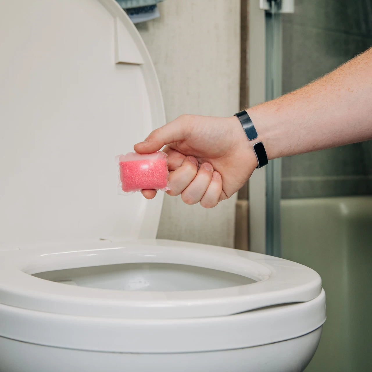 A hand holding a pink toilet cleaner gel packet above a white toilet in a bathroom setting.